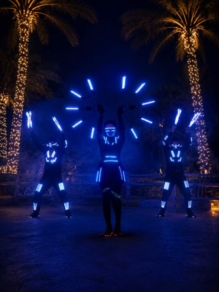 LED performers in blue illuminated costumes holding light props between palm trees at Al Ain Zoo.