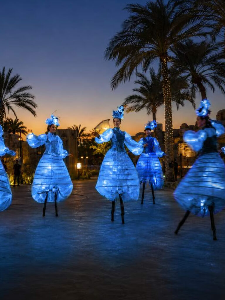 Stilt walkers in glowing blue costumes performing among palm trees at dusk at Al Ain Zoo.