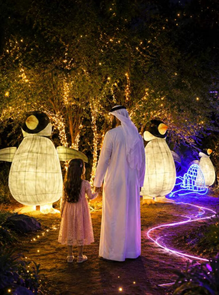 An adult and child beside illuminated penguin lanterns and a glowing garden path in Al Ain Zoo’s event imagery.